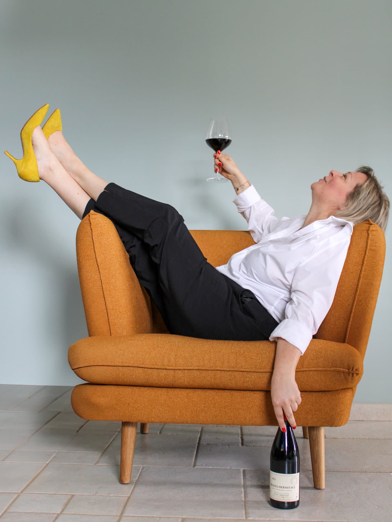 Sabrina Carlier allongée en travers d’un fauteuil orange, escarpins jaunes en l’air, levant un verre de vin rouge vers la lumière